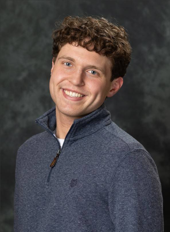 young man with big smile and brown hair wearing a navy blue quarter zip sweater and a grey background